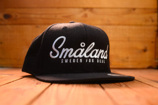 Black cap with 'Småland Sweden for Real' text on a wooden surface mangoebard