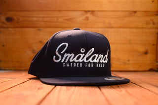 Navy cap with 'Småland Sweden for Real' text on a wooden surface and background from mangobeard