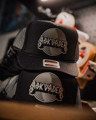Two black trucker hats stacked together, featuring a circular metallic silver and gold logo that reads 'ÅSKVÄDER' on the front panels and brim. The hats have black mesh side/back panels and a small white size tag on the brim.