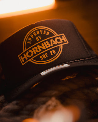 Close-up of a black Hornbach trucker cap with an orange ‘Approved by Hornbach SRF 26’ print, designed and pressed by Mangobeard for Sweden Rock Festival.