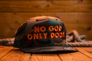 A camouflage snapback cap with orange embroidered text reading 'NO GOD ONLY DOOM' on the front, displayed on a wooden surface with a rope and wood plank background.